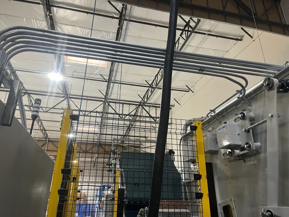Industrial machinery with overhead piping and safety barriers in a warehouse setting.